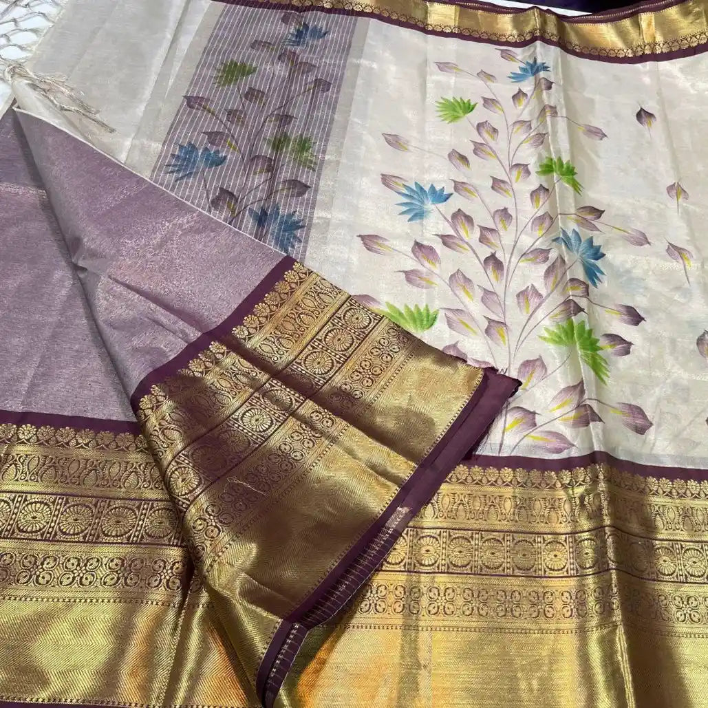 A brown and white soft Banarasi tissue silk saree by Swaya World. The saree has a white body with a hand-painted floral vine design in purple, blue, and green. It features a broad, contrasting golden Kanchipuram-style border and pallu. The pallu has tassels and is folded, showing the intricate golden weave and the reverse side of the hand-painted design.