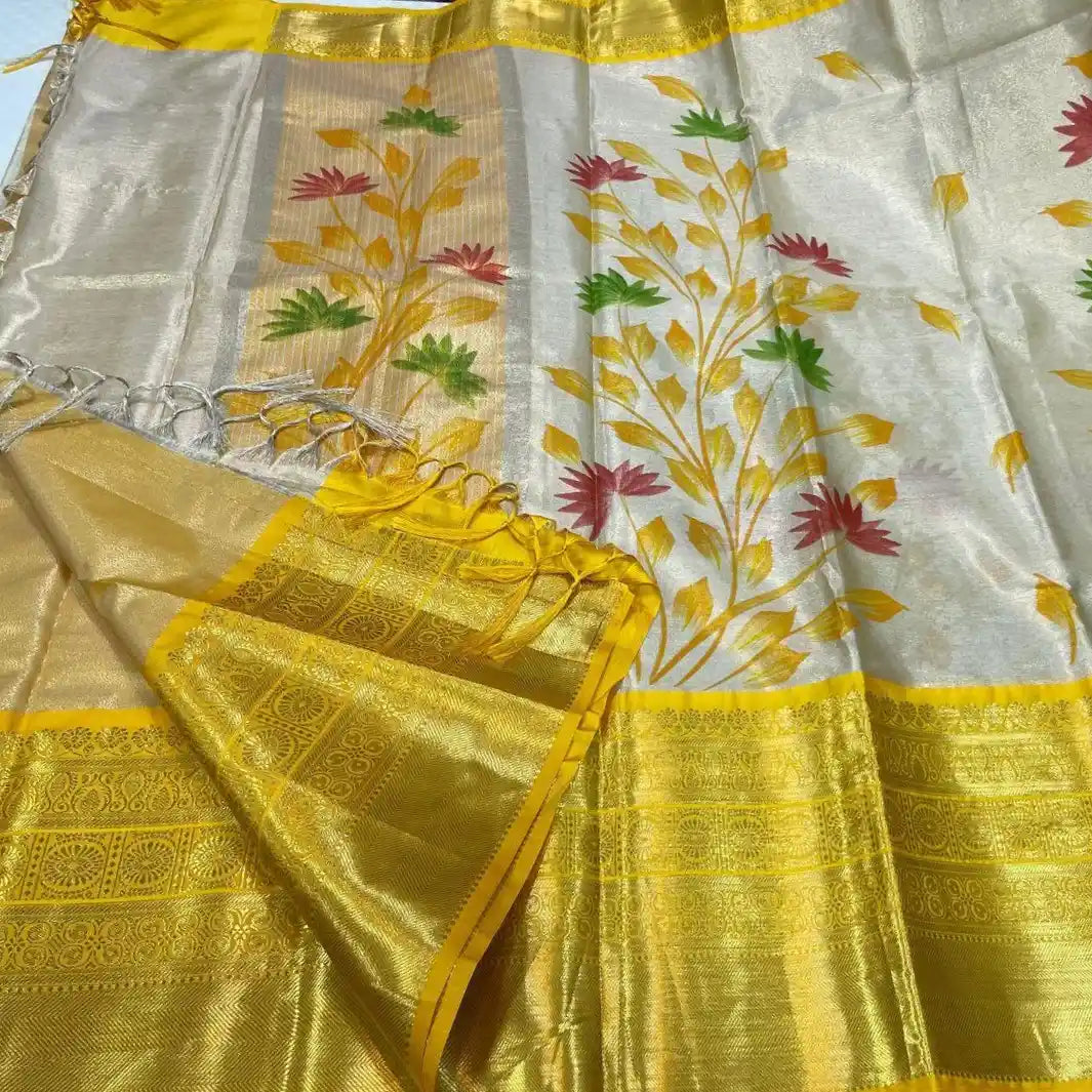 A yellow and white soft Banarasi tissue silk saree by Swaya World. The saree has a white body with a hand-painted floral vine design in yellow, red, and green. It features a broad, contrasting golden Kanchipuram-style border and pallu. The pallu has tassels and is folded, showing the intricate golden weave and the reverse side of the hand-painted design.